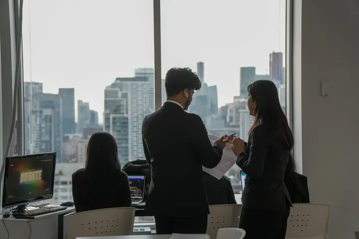 Two students standing at a window with the skyline behind them, one holding notes.