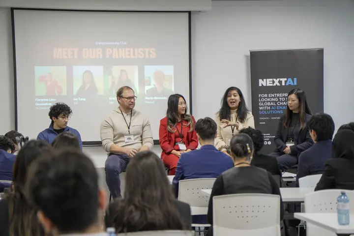 Panellists seated in front of a screen, a NEXT AI banner beside them, the audience in the foreground.