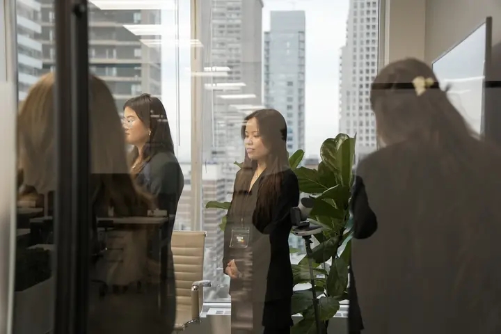 Seen through a glass wall: students standing in a meeting room beside a tall plant.