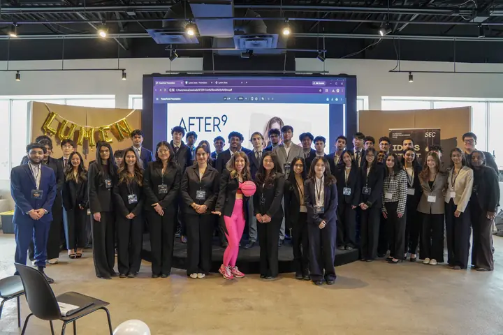 A large group of students posed in rows in front of a screen, gold LUMEN balloons at the left.