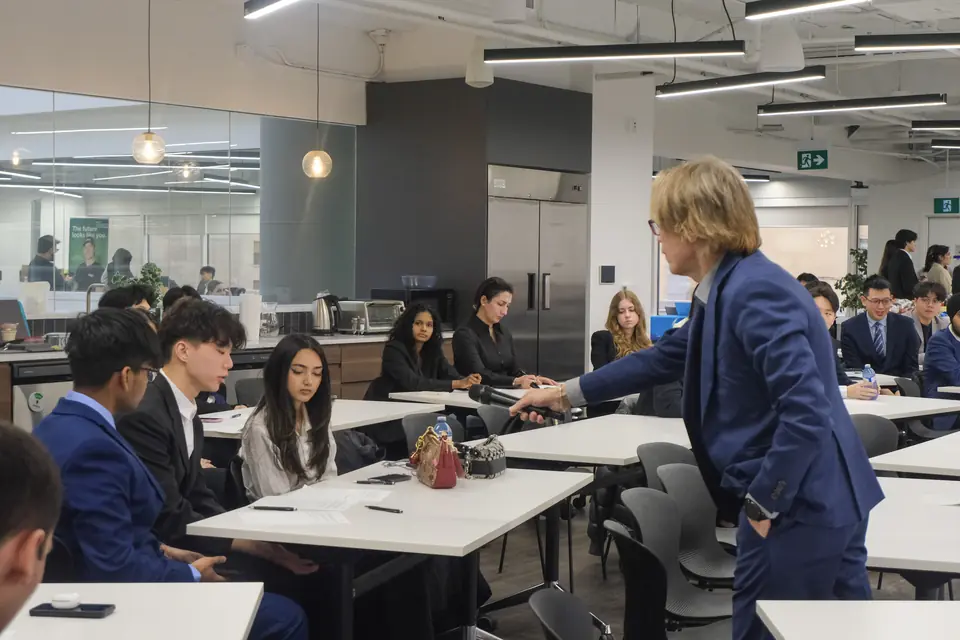 A man in a blue suit holds a microphone out to students seated at long white desks in an open-plan office.