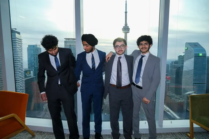 Four students in suits at a window with the CN Tower behind them.
